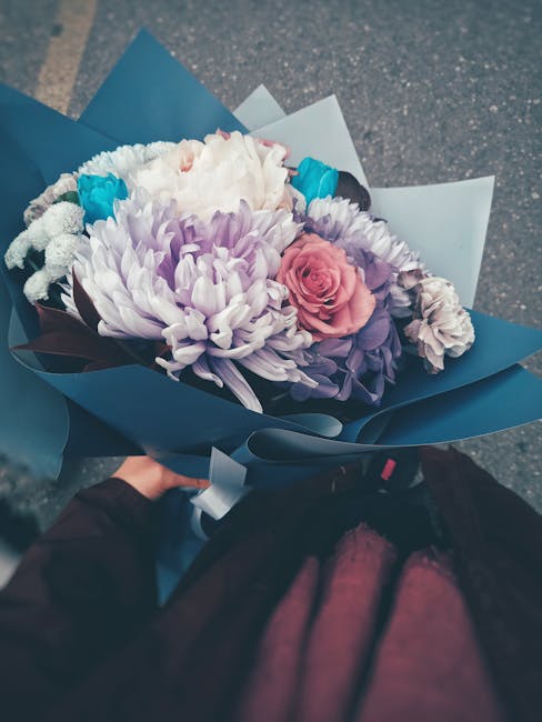 A hand holding a bouquet of fresh flowers wrapped in blue and white paper. The arrangement includes a pale pink rose, large white and purple chrysanthemums, and smaller filler flowers with purple and