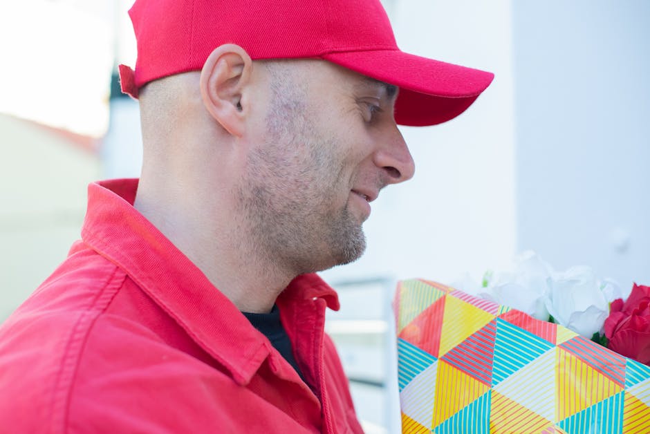 A man wearing a red baseball cap and a red jacket is holding a vibrant, geometrically patterned gift bag filled with fresh flowers. The bouquet features white and pink roses, arranged with green folia