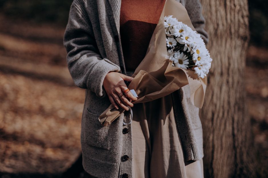 A person wearing a grey cardigan and beige trousers is holding a bouquet of fresh white daisies wrapped in light brown craft paper, with a beige satin ribbon, standing outdoors near a tree trunk. The 