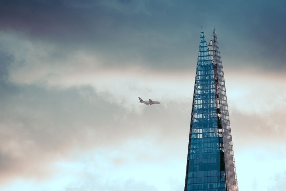 The image features the iconic The Shard skyscraper with its glass façade reflecting the cloudy sky, situated near London City Airport. In the background, an airplane is flying through the cloudy, ove