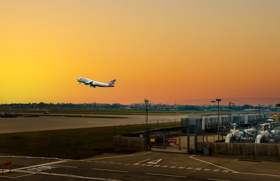 An airplane with British Airways livery is captured mid-takeoff, ascending against a vibrant orange and yellow sunset sky at London City Airport. The foreground features airport tarmac and ground serv
