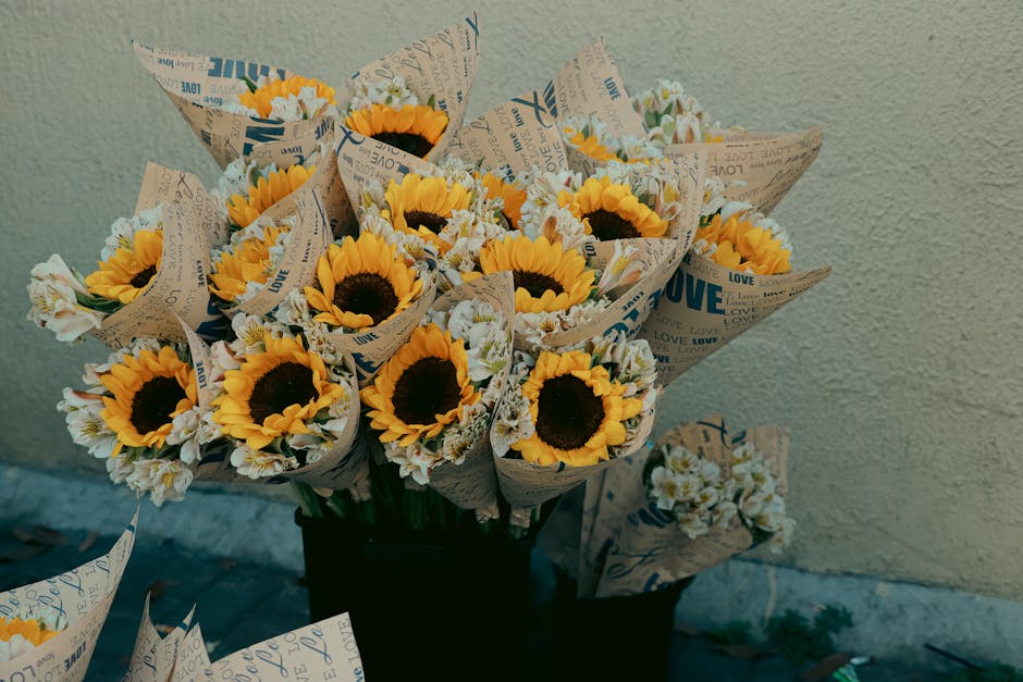 A vibrant bouquet of fresh yellow sunflowers with dark centers, accompanied by white and pale yellow filler flowers, all arranged in flower wrapping paper printed with words like 'LOVE' and 'LOVE' in 