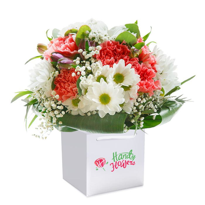 Carefully packaged in a sleek white gift bag, this vibrant bouquet seems to sing with the lively essence of Silvertown, its colours and textures suggesting both celebration and sentiment. Generous coral and peach carnations anchor the arrangement, their plush, ruffled petals conjuring memories of blooming gardens along the Thames Barrier Park. Peppered among them are luminous white daisies, each featuring a golden-green center that brings a burst of cheer reminiscent of sunlit walks through Lyle Park. The bouquet is softly punctuated by cascades of baby's breath, their cloud-like clusters adding a touch of innocence and lift. A scattering of partially closed, purple-tinged buds introduce an element of mystery, their compact form hinting at future blooms. Sweeping arcs of glossy, deep green foliage wrap the base and sides, their verdancy reflecting the renewal ongoing throughout Silvertown's parks and riverside spaces. The rounded, abundant shape of the bouquet is highlighted by balanced, even lighting-every petal and leaf clearly defined. Polished white cord handles rise at the bag's top, designed not only for graceful gifting but also as a symbol of neighbourly care, perhaps destined for a joyous occasion or heartfelt thank you in E16. This expertly crafted image exemplifies the freshness, generosity, and artistry behind local flower delivery in Silvertown-turning nature's beauty into a memorable, personal gift.